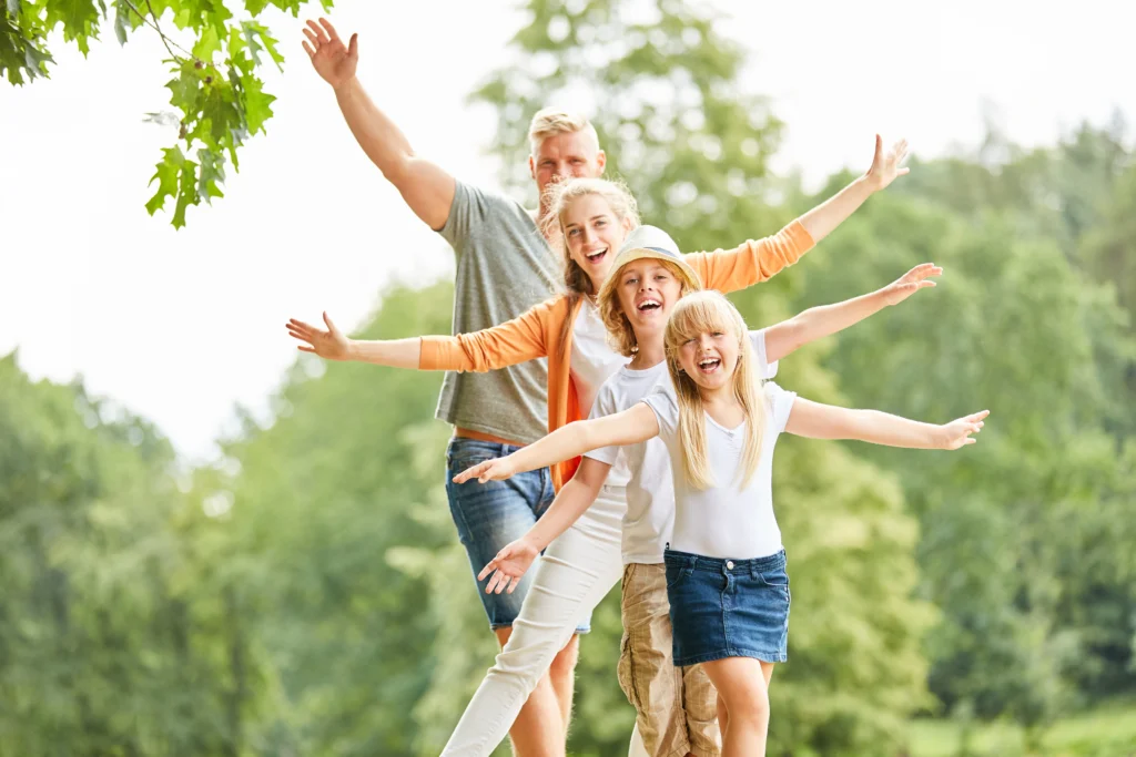 Eine fröhliche vierköpfige Familie steht im Freien und streckt lachend die Arme aus, um ihre Freude bei einem Ausflug zu zeigen.