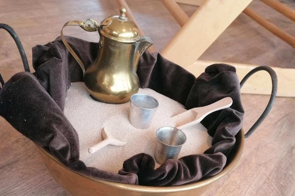 Eine Metallschüssel gefüllt mit Sand, darin eine goldene Kanne, kleine Metallbecher und Holzwerkzeuge für spielerische Aktivitäten.