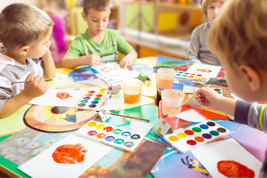 Eine Gruppe von Kindern sitzt an einem bunten Tisch und malt konzentriert mit Wasserfarben in einem Familienzentrum.