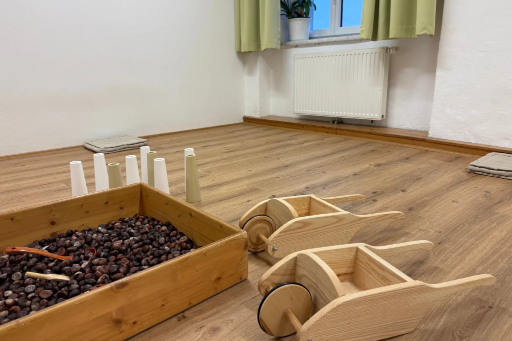 Spielbereich im Familienzentrum mit einer Holzkiste voller Kastanien, zwei kleinen Holzschubkarren und weißen Kegeln auf dem Boden.