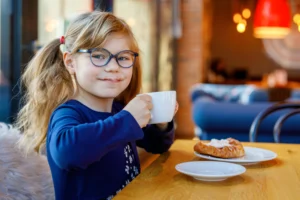 Ein kleines Mädchen mit Brille trinkt in einem gemütlichen Café aus einer weißen Tasse und isst ein Gebäckstück.