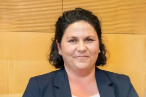 Eine freundlich lächelnde Frau mit dunklen Haaren vor einem hellen Holzhintergrund, Porträtaufnahme für das Familienzentrum.