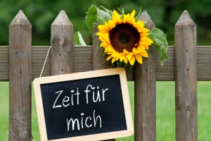 Eine Tafel mit der Aufschrift Zeit für mich hängt an einem Holzzaun, geschmückt mit einer leuchtend gelben Sonnenblume.