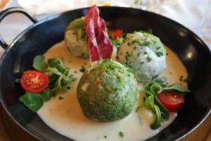 Drei grüne Spinatknödel in einer hellen Käsesoße, garniert mit frischen Tomaten und Salat in einer schwarzen Pfanne serviert.