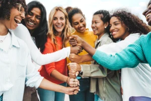Eine diverse Gruppe fröhlicher Frauen hält gemeinsam die Fäuste in die Mitte als Symbol für Zusammenhalt und Gemeinschaft.