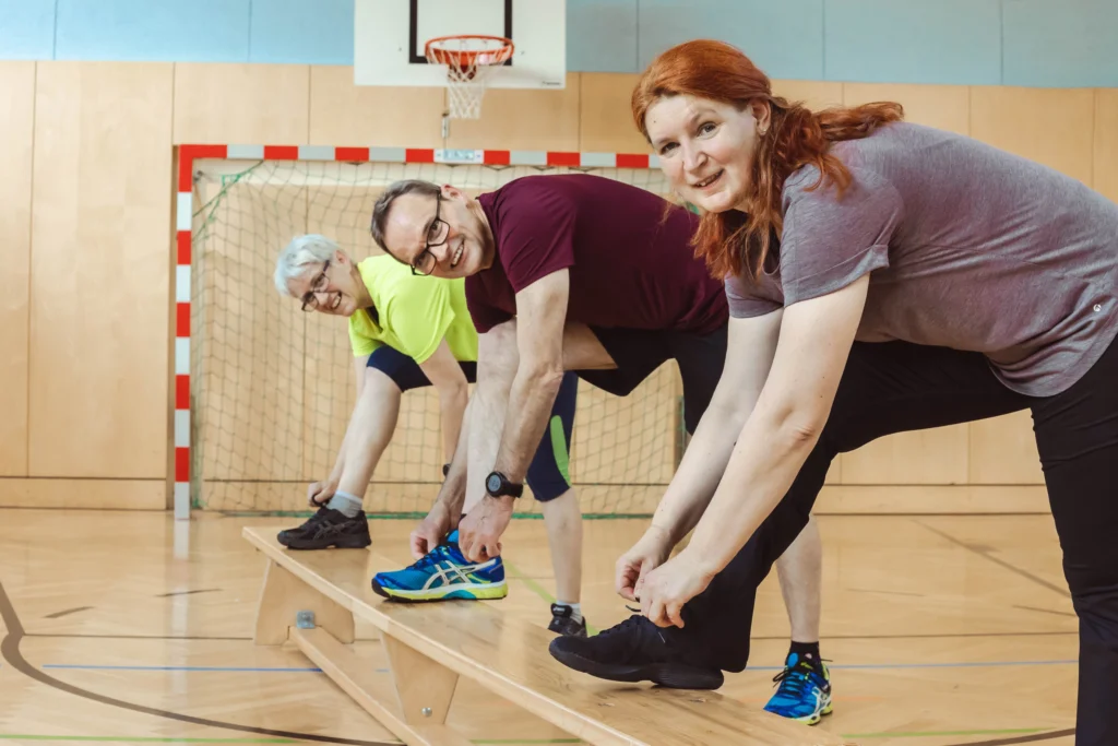 Drei Personen in Sportkleidung binden sich in einer Turnhalle die Schnürsenkel ihrer Sportschuhe auf einer Holzbank.