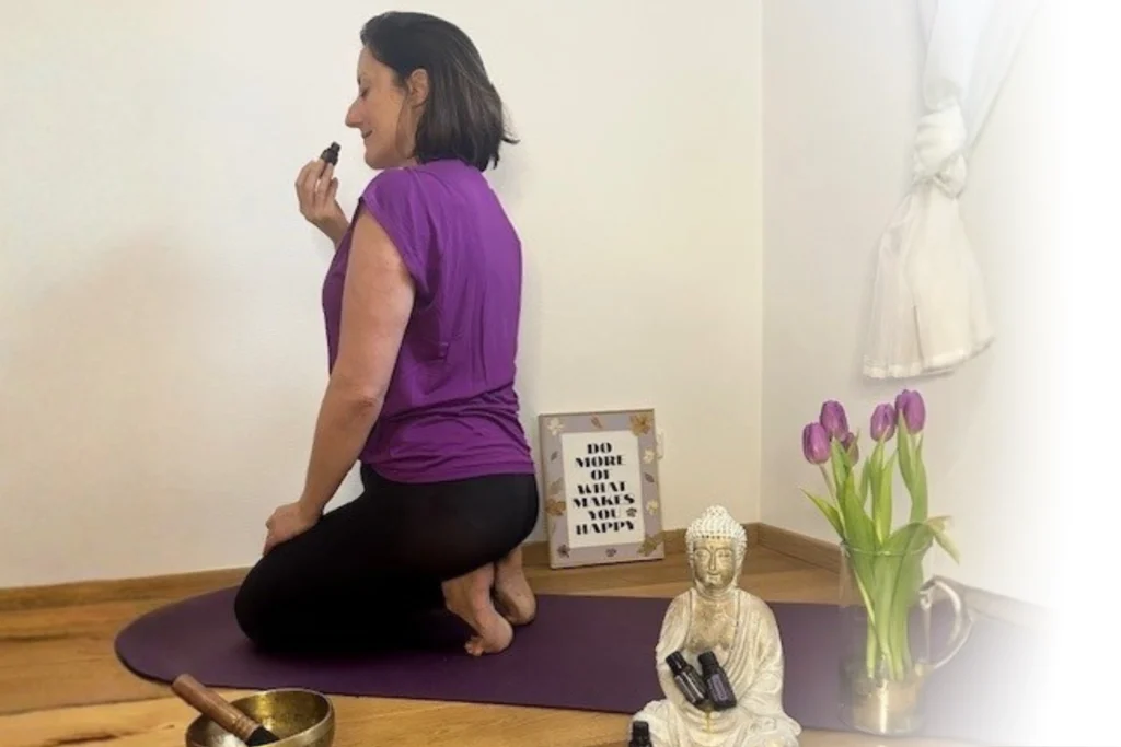 Eine Frau in einem lila Oberteil kniet auf einer Yogamatte und schnuppert an einem ätherischen Öl in einer entspannten Umgebung.