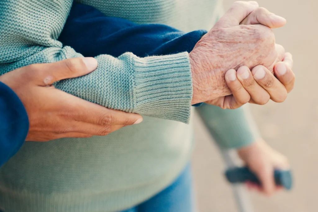 Eine helfende Hand hält sanft die Hand einer älteren Person, die sich auf einen Gehstock stützt, als Symbol für Unterstützung.