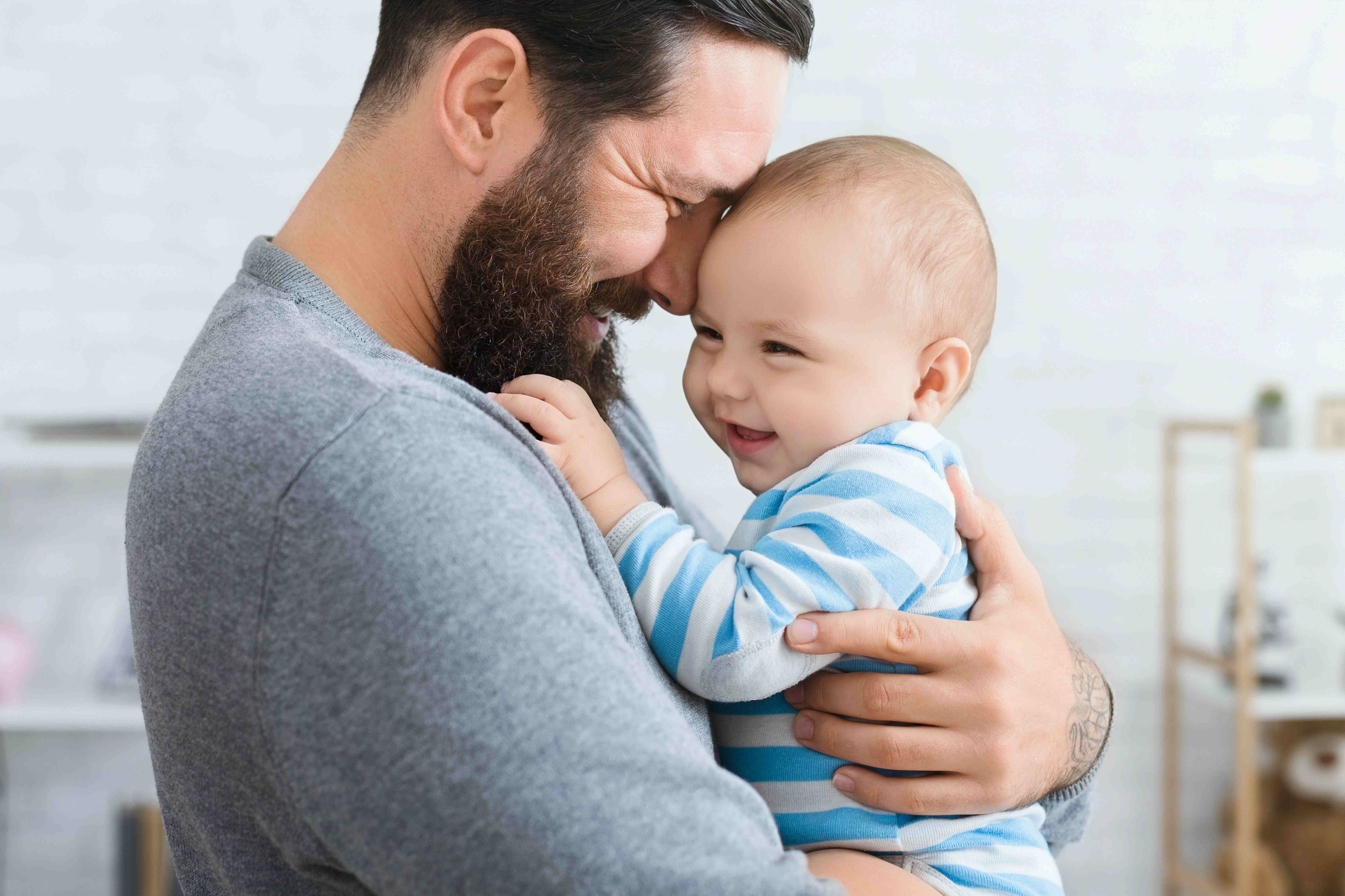 Ein junger Mann hält ein Baby mit blau-weiß gestreiften Pullover auf dem Arm. Beide lächeln.