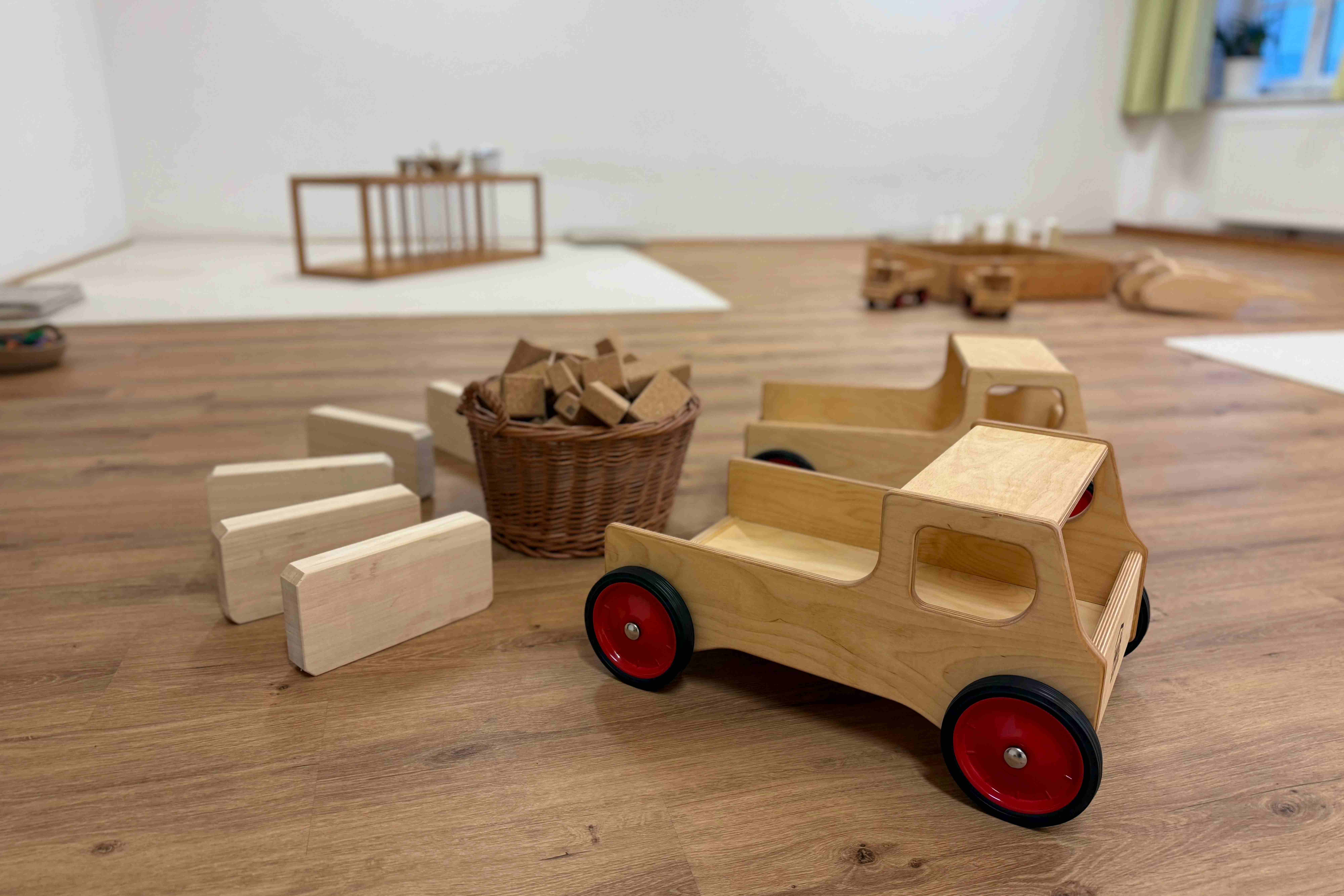 Spielbereich im Familienzentrum mit Holzautos, Bausteinen und einem Weidenkorb auf einem hellen Holzboden für Kinder.