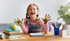 Ein fröhliches Mädchen mit bemalten Händen sitzt am Tisch und freut sich über kreative Bastelaktivitäten im Familienzentrum.