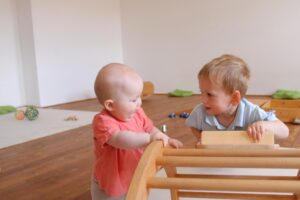 Ein Kleinkind und ein Baby spielen gemeinsam an einem hölzernen Kletterbogen in einem hellen Gruppenraum des Familienzentrums.
