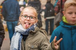 Ein fröhliches Mädchen mit Brille und Schal streckt bei einer Familienveranstaltung im Freien die Zunge heraus und zeigt ein V-Zeichen.