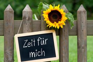 Ein Gartenzaun aus Holz an dem eine Sonnenblume und ein Schild zu sehen sind. Am Schild steht Zeit für mich mit Kreise auf eine schwarze Tafel geschrieben.
