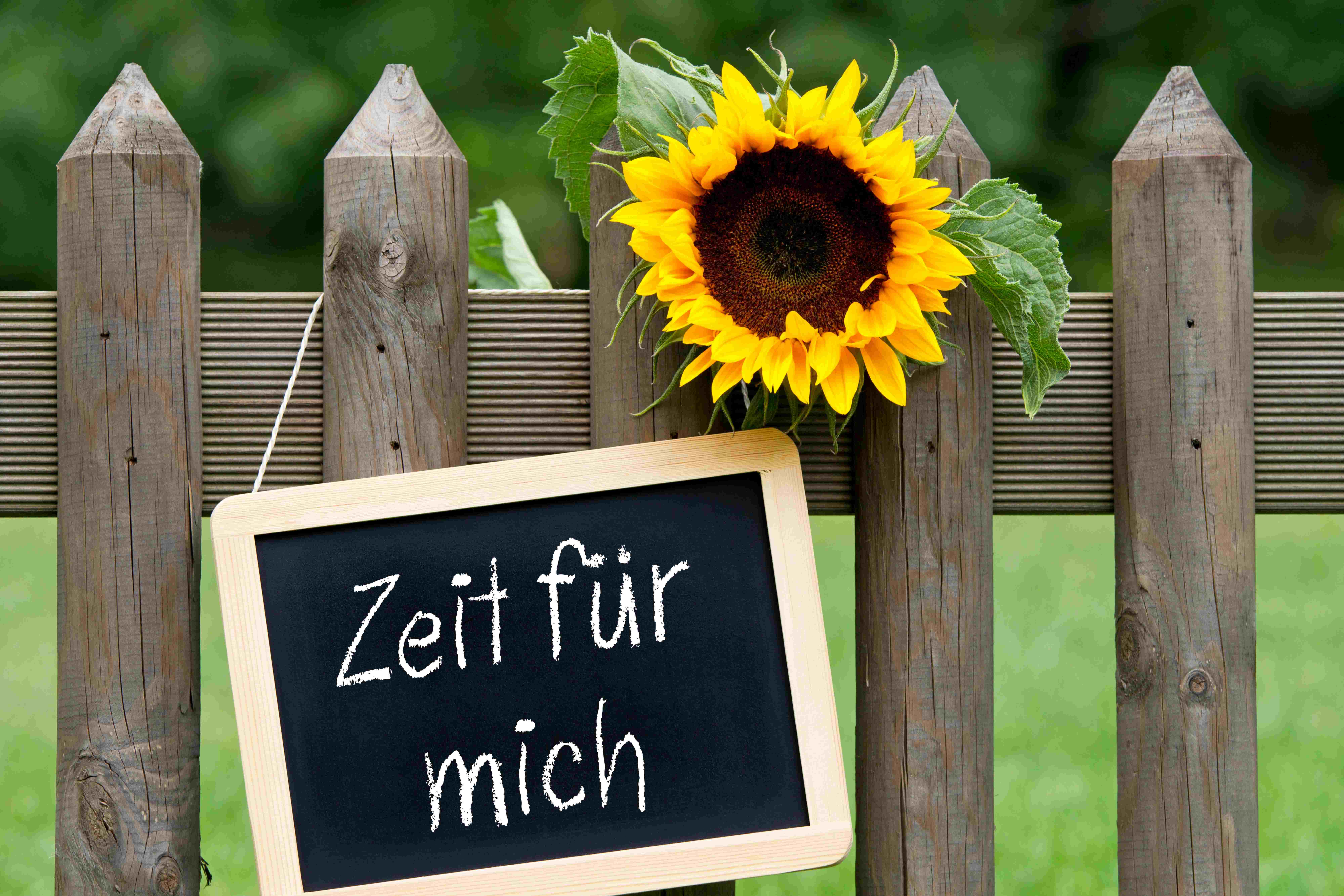 Ein Gartenzaun aus Holz an dem eine Sonnenblume und ein Schild zu sehen sind. Am Schild steht Zeit für mich mit Kreise auf eine schwarze Tafel geschrieben.