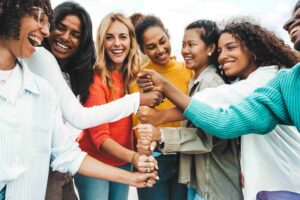 Eine diverse Gruppe fröhlicher Frauen hält gemeinsam die Fäuste in die Mitte als Symbol für Zusammenhalt und Gemeinschaft.