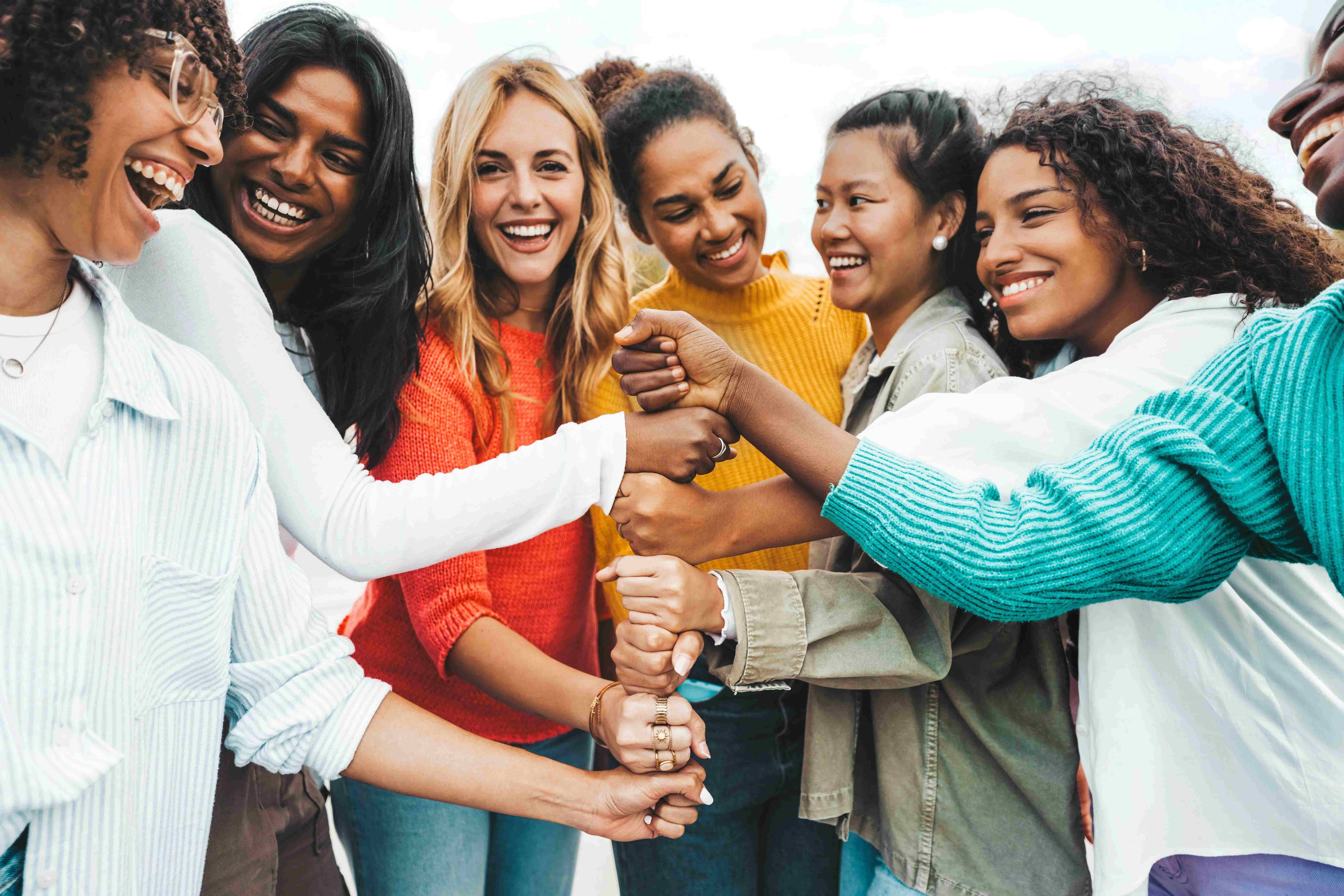 Eine diverse Gruppe fröhlicher Frauen hält gemeinsam die Fäuste in die Mitte als Symbol für Zusammenhalt und Gemeinschaft.