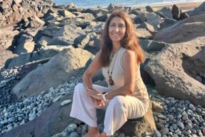 Eine lächelnde Frau in heller Kleidung sitzt entspannt auf einem großen Felsen an einem steinigen Strand am Meer.