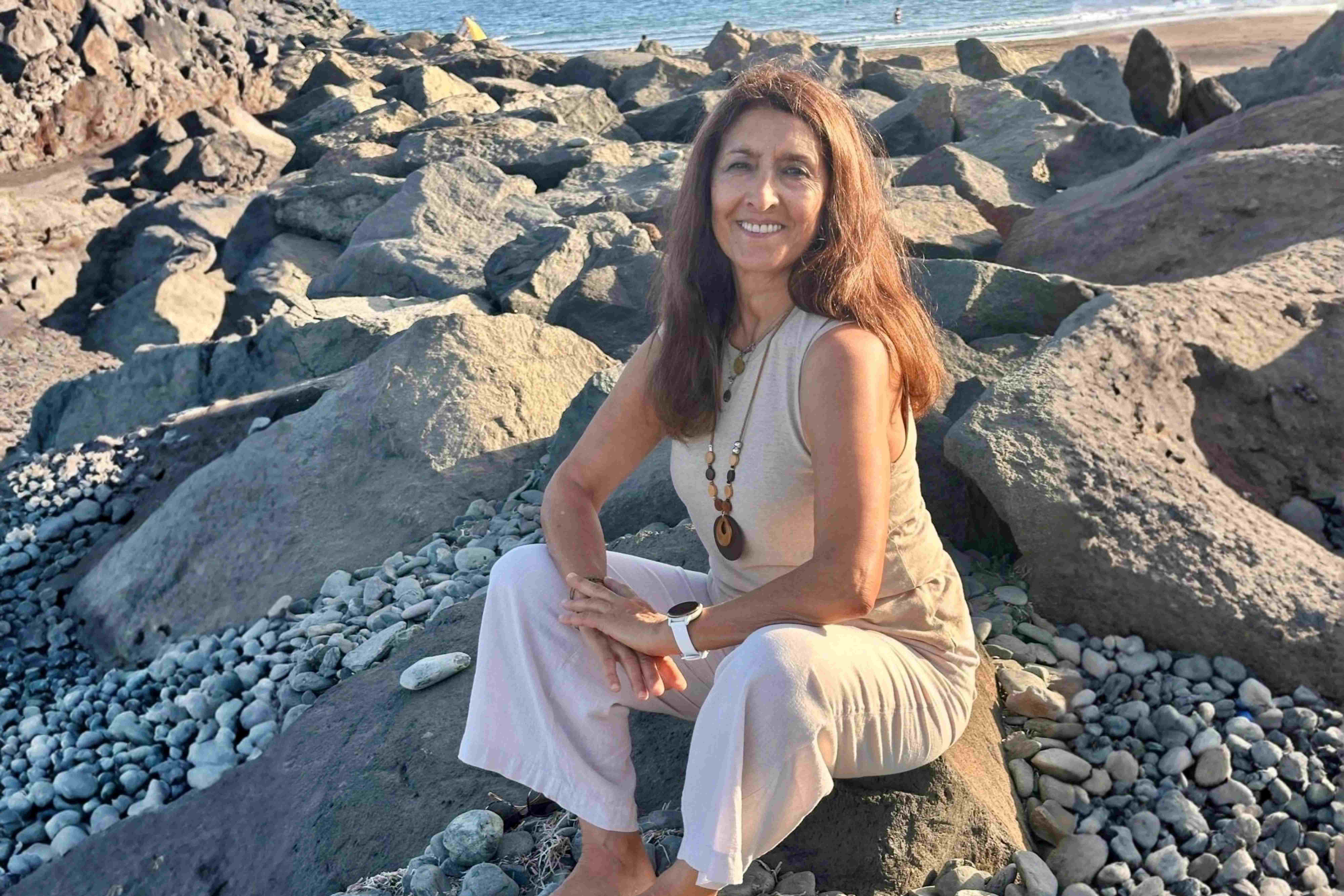 Eine lächelnde Frau in heller Kleidung sitzt entspannt auf einem großen Felsen an einem steinigen Strand am Meer.