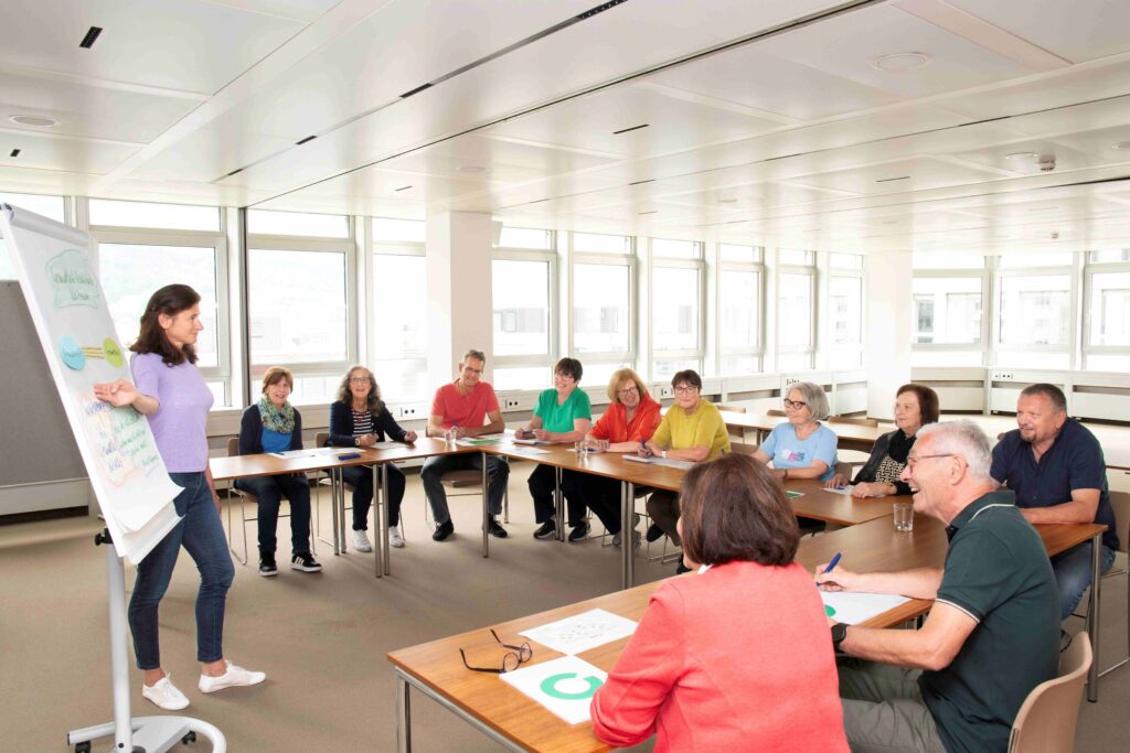 Eine Kursleiterin präsentiert vor einer Gruppe Erwachsener an einem Flipchart in einem hellen, modernen Seminarraum.