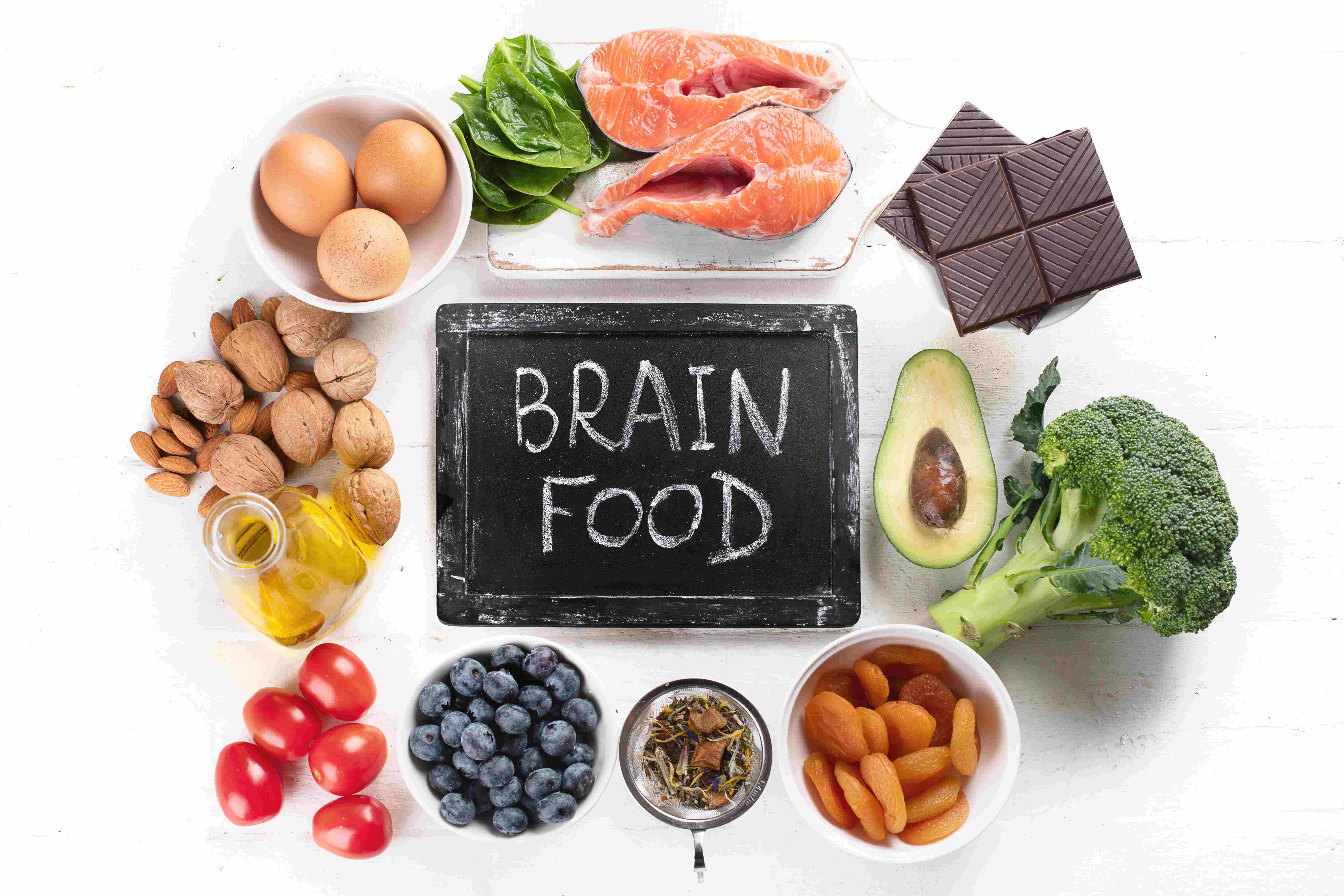 Eine Auswahl an gesundem Brainfood wie Lachs, Nüsse, Beeren, Gemüse und dunkle Schokolade auf einem weißen Holztisch arrangiert.