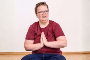 Eine Person mit Brille und weinrotem T-Shirt sitzt auf dem Boden und hält die Hände in einer achtsamen Geste vor der Brust.